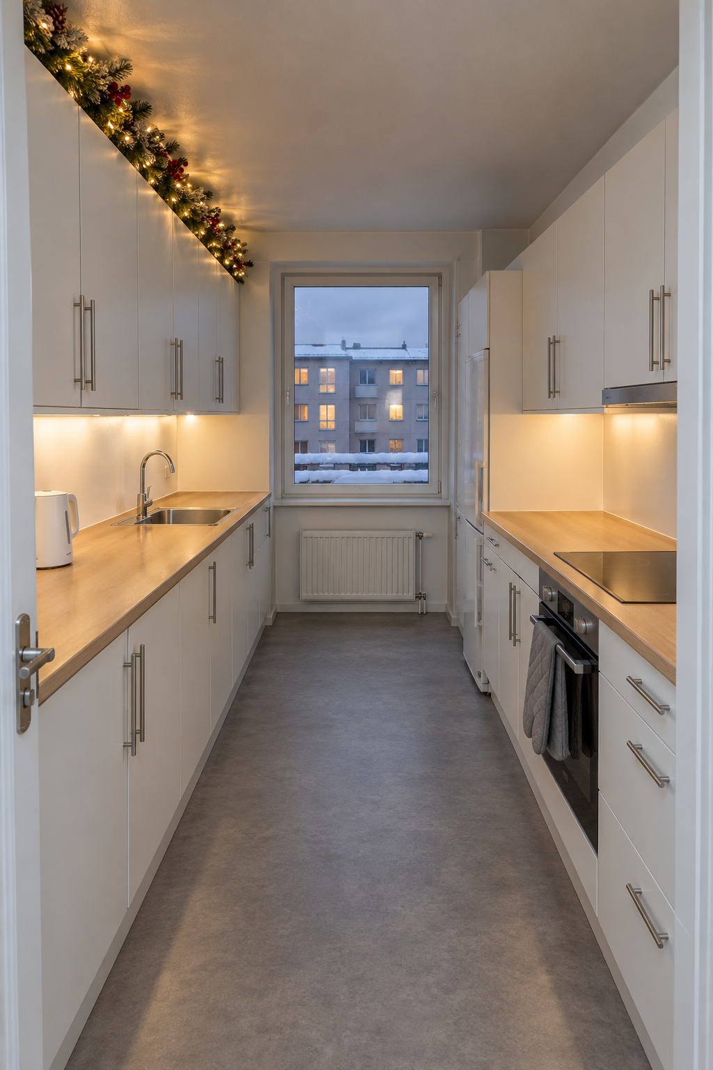 Small apartment galley Christmas kitchen with a lit garland with red berries and frosted pinecones along the top of flat white upper cabinets and clear counters